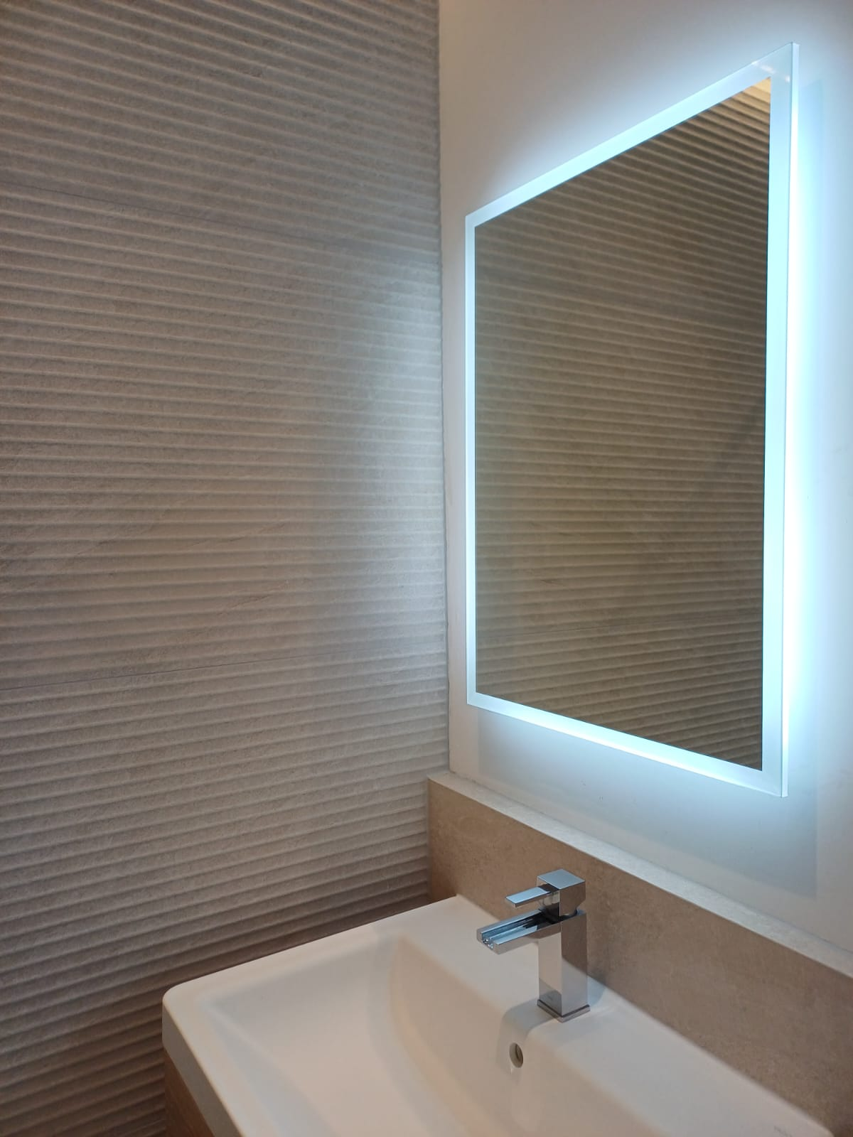 Illuminated LED bathroom mirror above a basin with textured stone-effect wall tiles